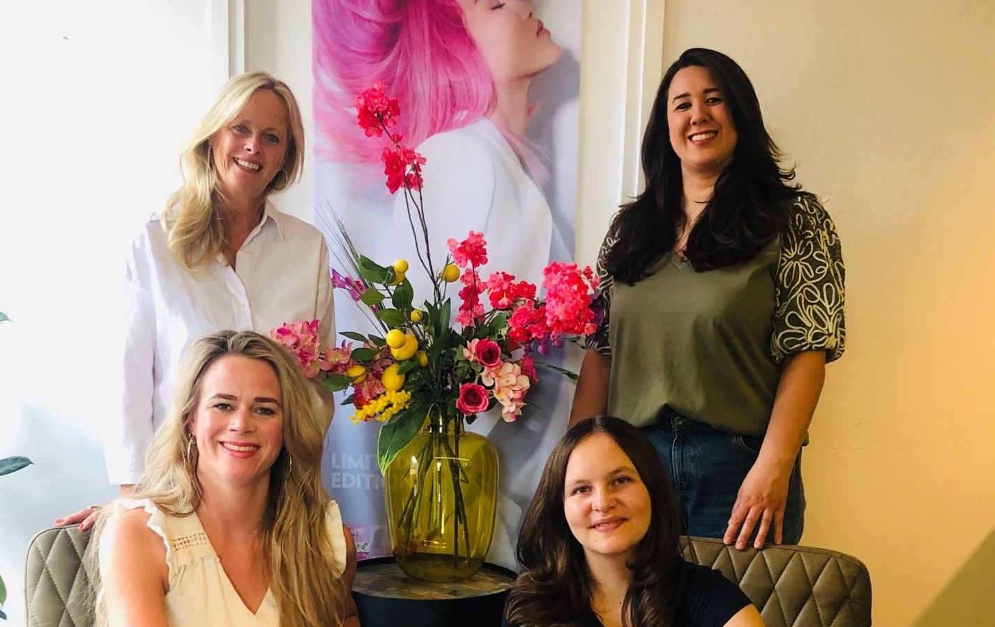 Het team van Koele Coupe in de salon: vier kapsters bij een boeket bloemen en een poster met roze haar.